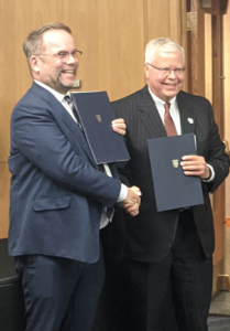 Hessen State Minister of Science and Research, Arts and Culture, Timon Gremmels stands next to Universities of Wisconsin President Jay O. Rothman shaking hands and smiling at the camera while both hold copies of the Letter of Intent.