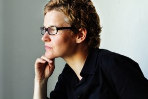 A woman in glasses and a black button-down looks to the right.